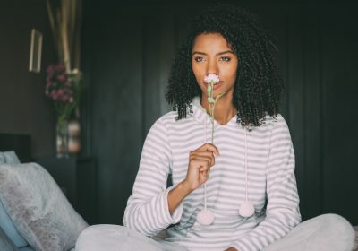 close up of a pretty black woman with curly hair smelling a rose flower sit on bed looking away