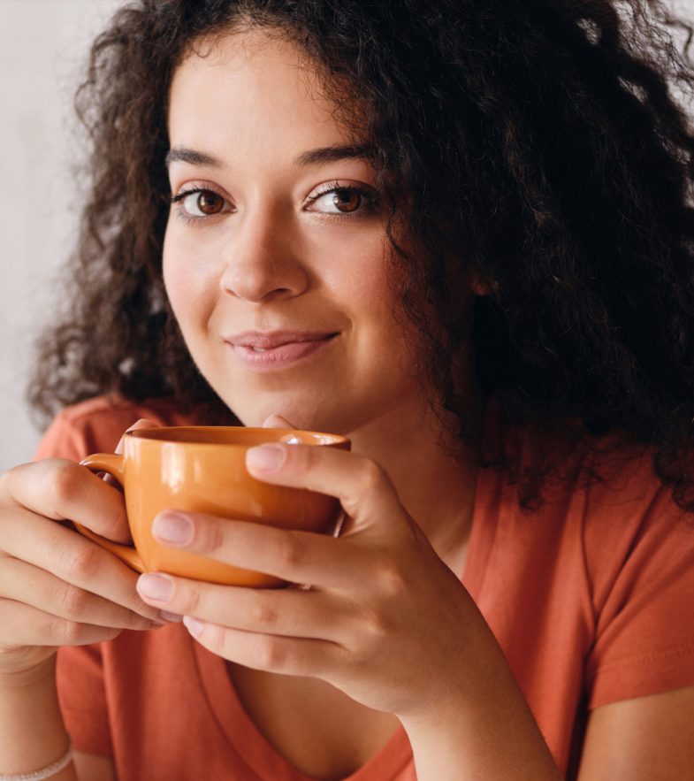 Portrait of young beautiful smiling woman with dark curly hair holding orange cup of coffee in hands dreamily looking in camera at modern cozy home alone